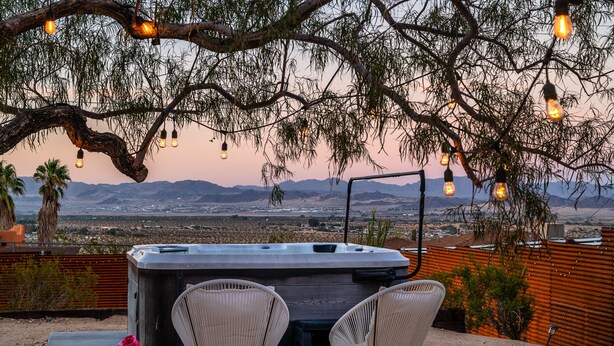A hot tub with two chairs, surrounded by a wooden fence, overlooking a desert landscape with mountains in the distance. - ★Hot tub under the stars★ 3 King Beds, Cowboy Pool, Desert Views, Fire Pit, EV (Twentynine Palms)