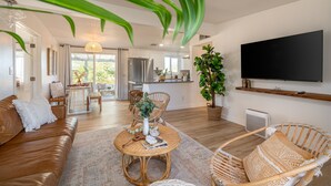 A modern living room with a brown leather sofa, wicker chairs, a round coffee table, and a large flat-screen TV mounted on the wall. - ★Hot tub under the stars★ 3 King Beds, Cowboy Pool, Desert Views, Fire Pit, EV (Twentynine Palms)