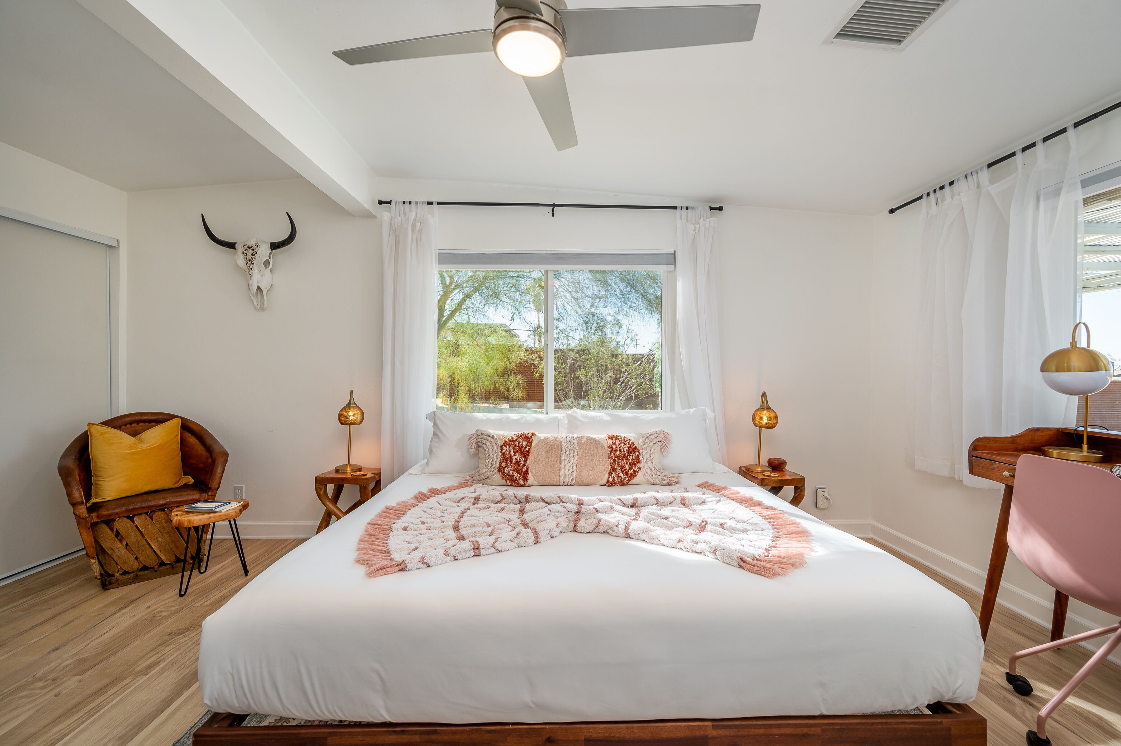A bedroom with a large bed, a ceiling fan, a window with curtains, a desk with a lamp, and a chair.