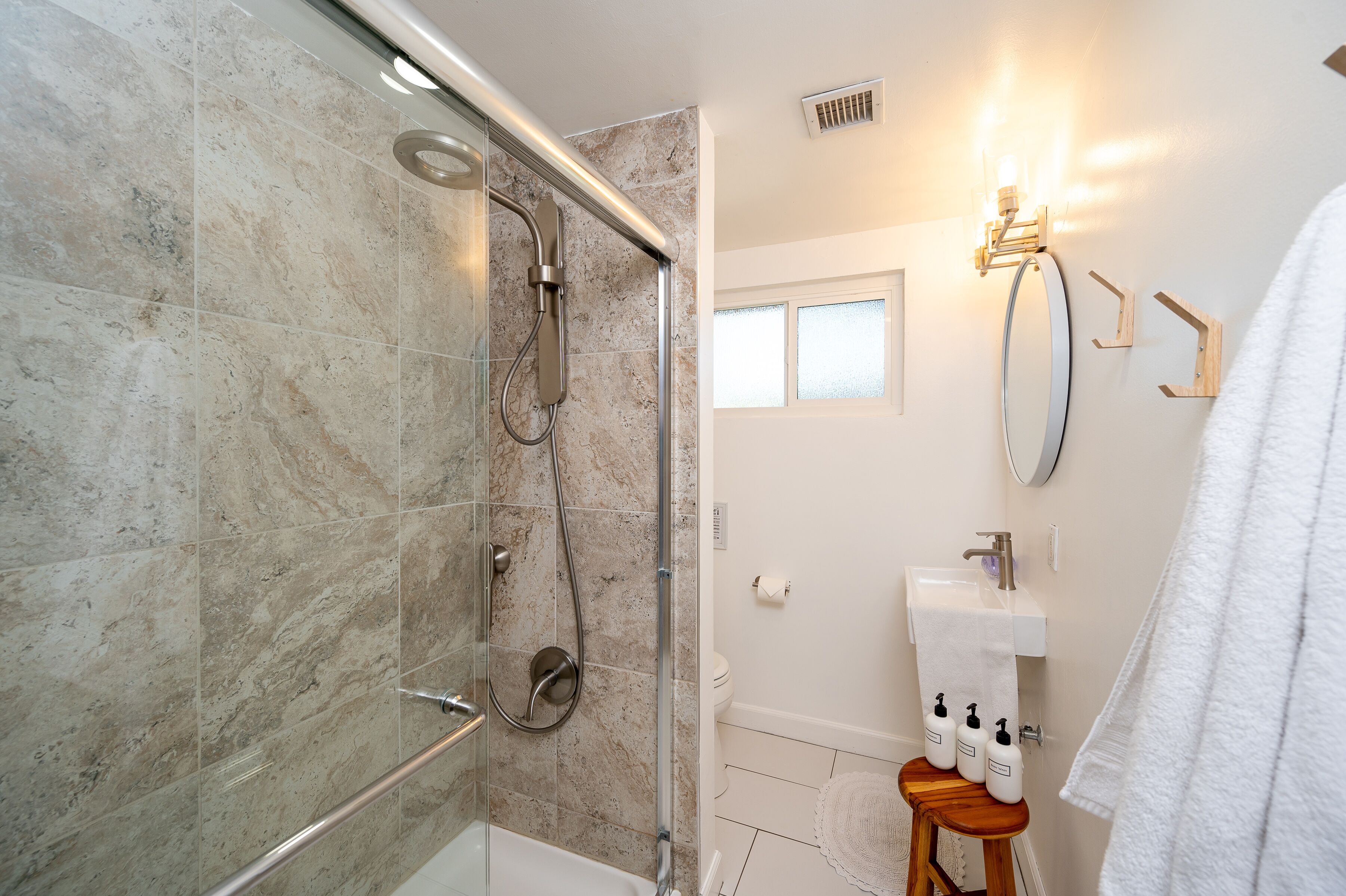Une salle de bain moderne avec une douche à parois de verre, un lavabo blanc avec un miroir et un tabouret en bois sur lequel sont posés des articles de toilette.