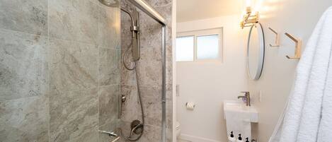 Une salle de bain moderne avec une douche à parois de verre, un lavabo blanc avec un miroir et un tabouret en bois sur lequel sont posés des articles de toilette.