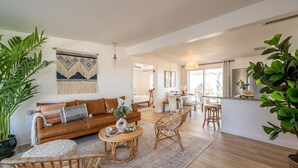 A spacious living area with a brown leather sofa, wicker chairs, a round coffee table, and a large potted plant. - ★Hot tub under the stars★ 3 King Beds, Cowboy Pool, Desert Views, Fire Pit, EV (Twentynine Palms)