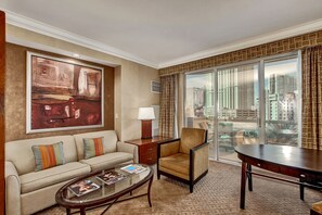 Quarto de hotel com uma janela grande, um sofá, uma poltrona, uma mesinha e um quadro na parede.
