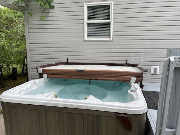 Un jacuzzi con borde de madera y parte interior blanca, ubicado al aire libre frente a una casa con una ventana y un enchufe a la vista.