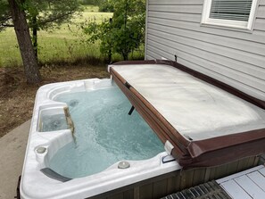 Un bain à remous doté d’un couvercle en bois, installé à l’extérieur, à côté d’une maison avec une fenêtre.
