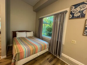 A bedroom with a bed, a window with curtains, a painting on the wall, and a wooden floor.