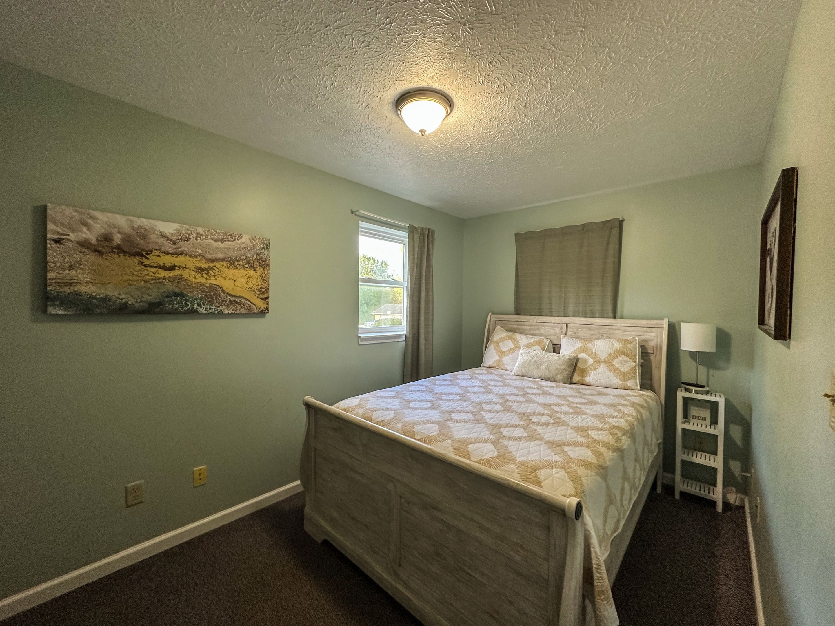 Un dormitorio con una cama, una mesita de noche, una lámpara, una ventana con cortinas y un cuadro en la pared.