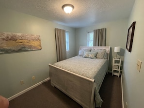 Quarto com cama, mesa de cabeceira, abajur e um quadro na parede.