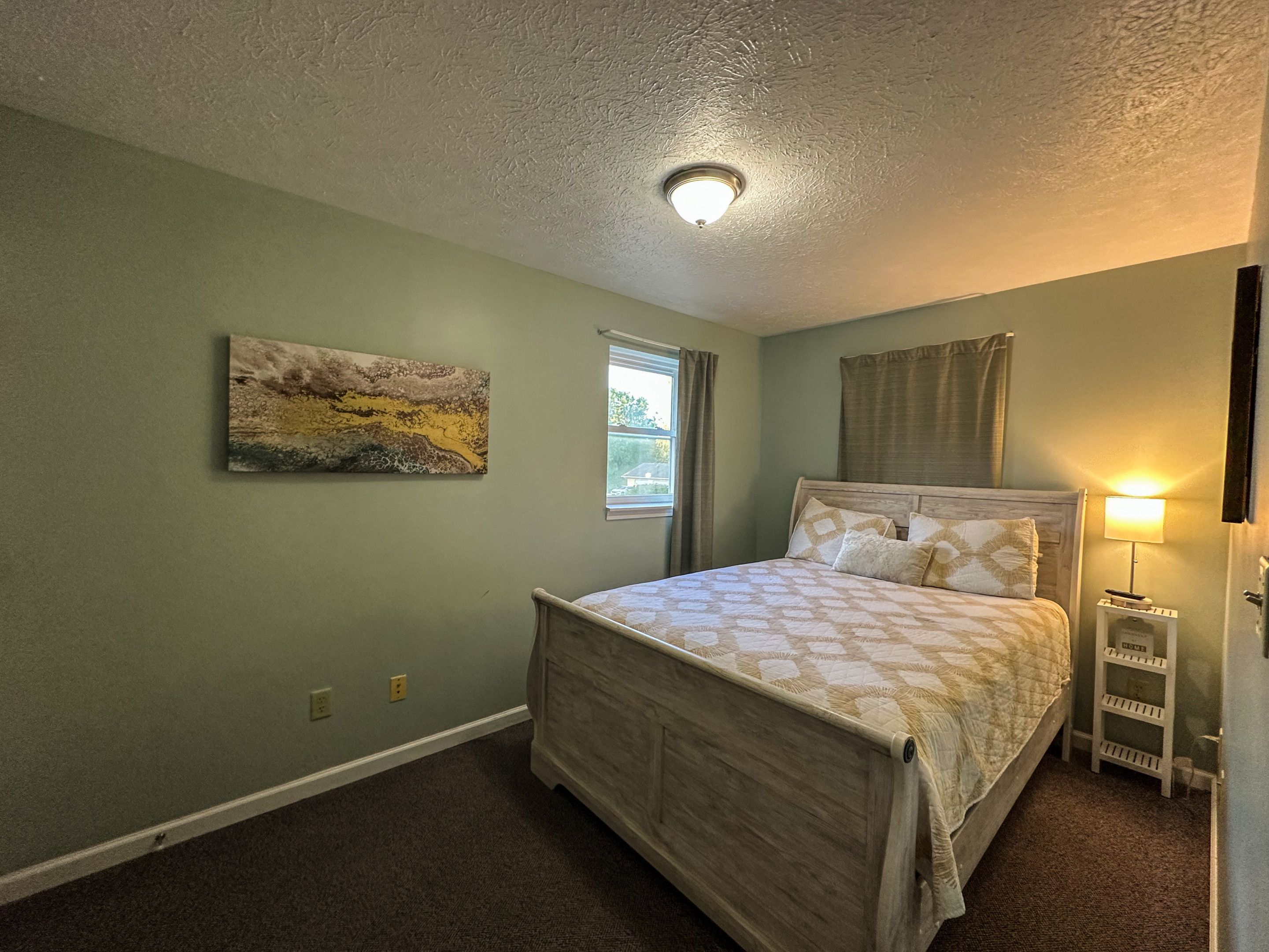 A bedroom with a bed, a window, a painting, and a nightstand with a lamp.