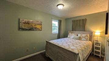 A bedroom with a bed, a window, a painting, and a nightstand with a lamp.