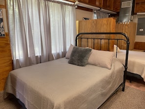 Una cama individual con estructura metálica, almohada gris y colcha color beige en una habitación con paredes de madera y una ventana con cortinas traslúcidas.