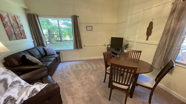 Ein Zimmer mit braun-lederner Couch, einem kleinen Tisch mit Stühlen, einem Fernseher und einem Fenster mit Vorhängen.