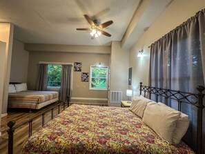 Une chambre à coucher avec un grand lit, un plus petit lit, un ventilateur de plafond, une fenêtre avec des rideaux et une table de chevet avec une lampe.