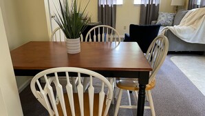 A dining area with a wooden table and white chairs, a potted plant, and a grey sofa in the background.