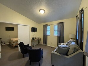 A compact living room with a sofa, a chair, a TV, a desk, and a bed.