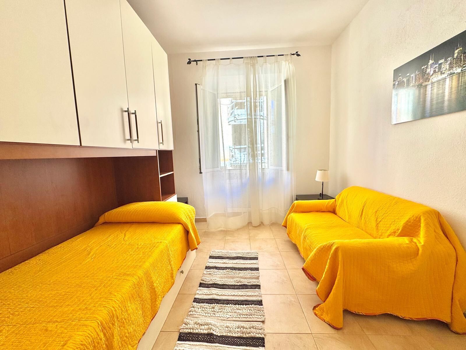 A room with two beds, a window with sheer curtains, a lamp, and a cityscape painting.