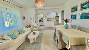 A modern living room with a sofa, coffee table, and dining area. There is a kitchen in the background with white cabinets and a view of the sea.