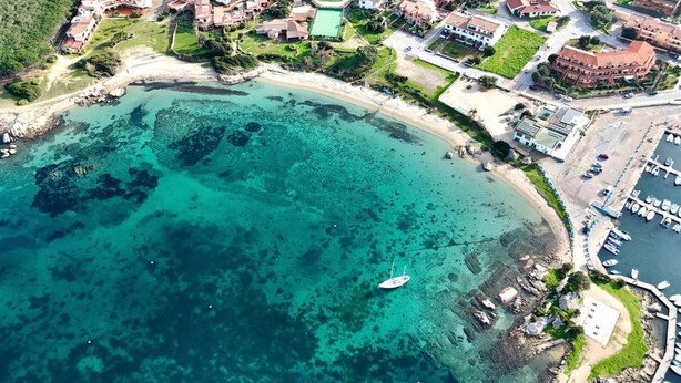 An aerial view of a coastal area with a marina, buildings, and clear turquoise waters.