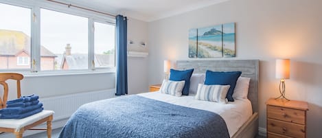 A bedroom with a bed, a chair with folded towels, a nightstand, a lamp, and a window with curtains.