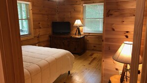 A wooden cabin room with a bed, a television on a wooden cabinet, and a lamp on a wooden stool. - SUMMER SPECIAL — Book 5 nights 6+7th FREE — LOG CABIN with Privacy+ Firepit (West Jefferson)