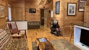 A cozy cabin interior with wooden walls, a sofa, a bench, a fireplace, and framed pictures on the walls. - SUMMER SPECIAL — Book 5 nights 6+7th FREE — LOG CABIN with Privacy+ Firepit (West Jefferson)