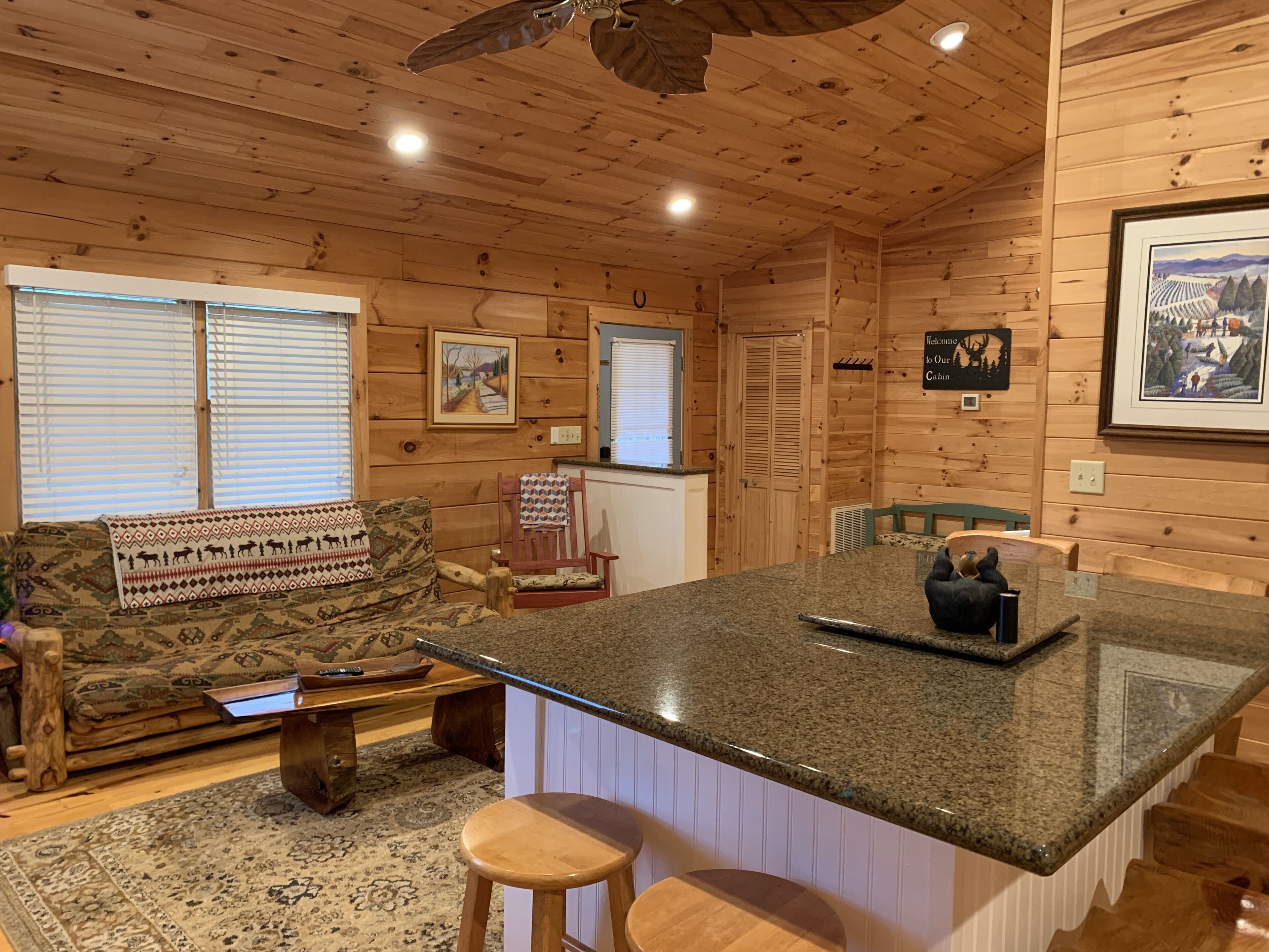 A cabin interior with a wooden ceiling, a kitchen island, a sofa with patterned cushions, and a rug.