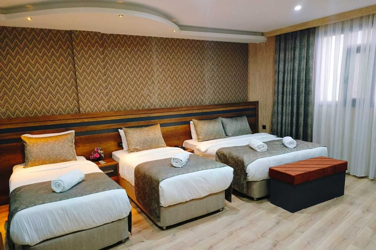 Ein Hotelzimmer mit drei Einzelbetten, einem Holzkopfstück, einem großen Fenster mit Vorhängen und einer Wand mit Muster.