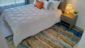 A neatly made bed with a white duvet, a tufted headboard, and a bedside table with a lamp and a small plant. - Astoria Apartments (Nairobi)