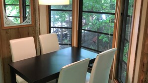 A dining area with a dark wooden table and white chairs, large windows with curtains, and a hanging light fixture.