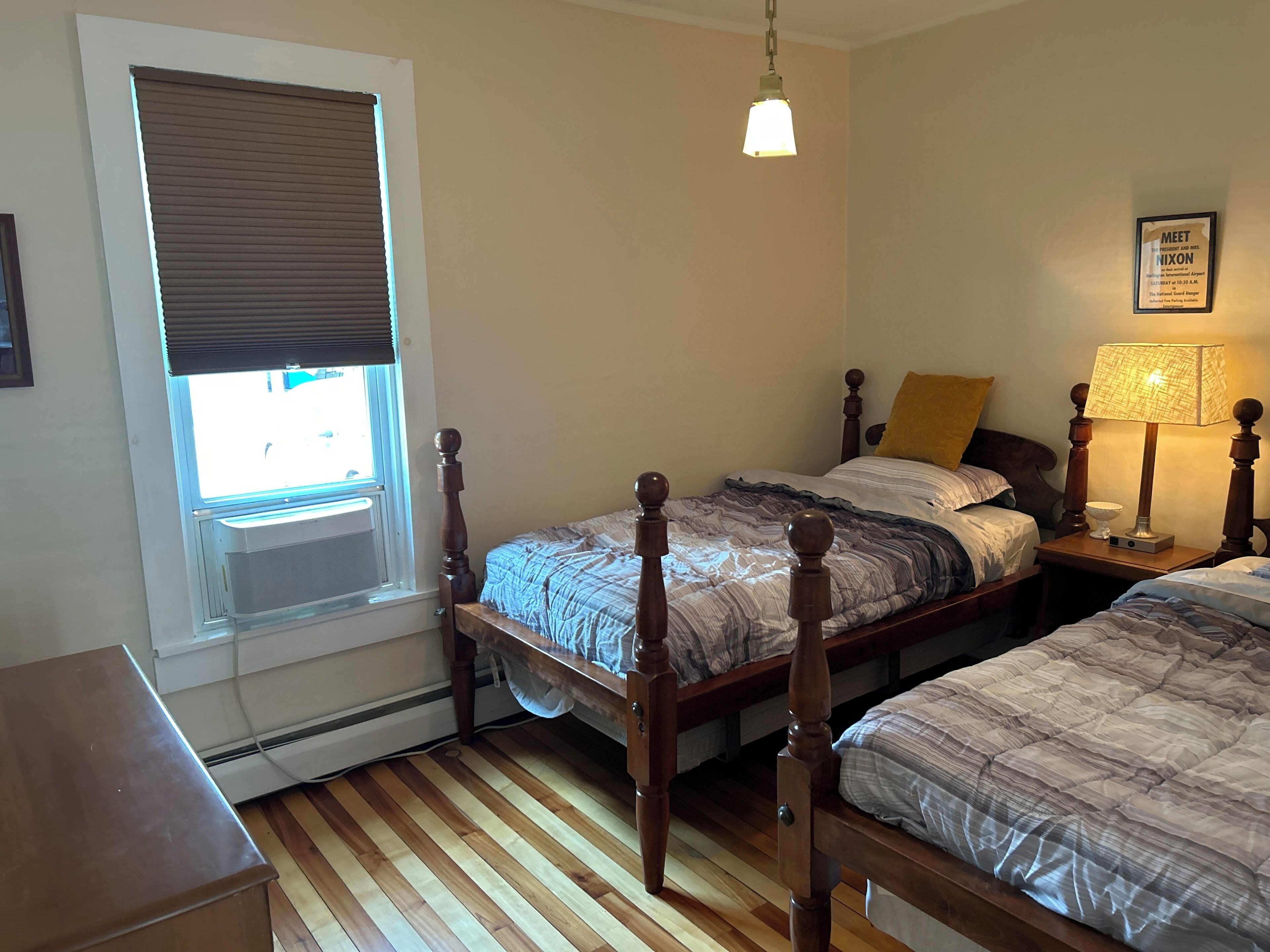 A bedroom with two wooden beds, a window with blinds, a hanging light fixture, and a framed sign on the wall.