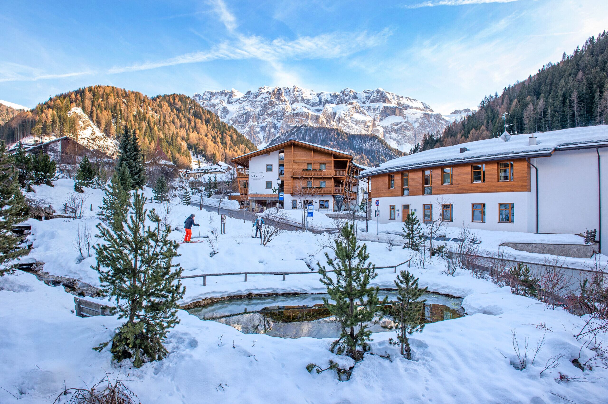 A snow-covered landscape with a wooden building, a small stream, and mountains in the background.