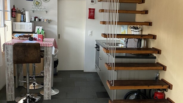 A modern kitchen with a wooden staircase, a dining area with a red and white checkered tablecloth, and a kitchen island with a round table.