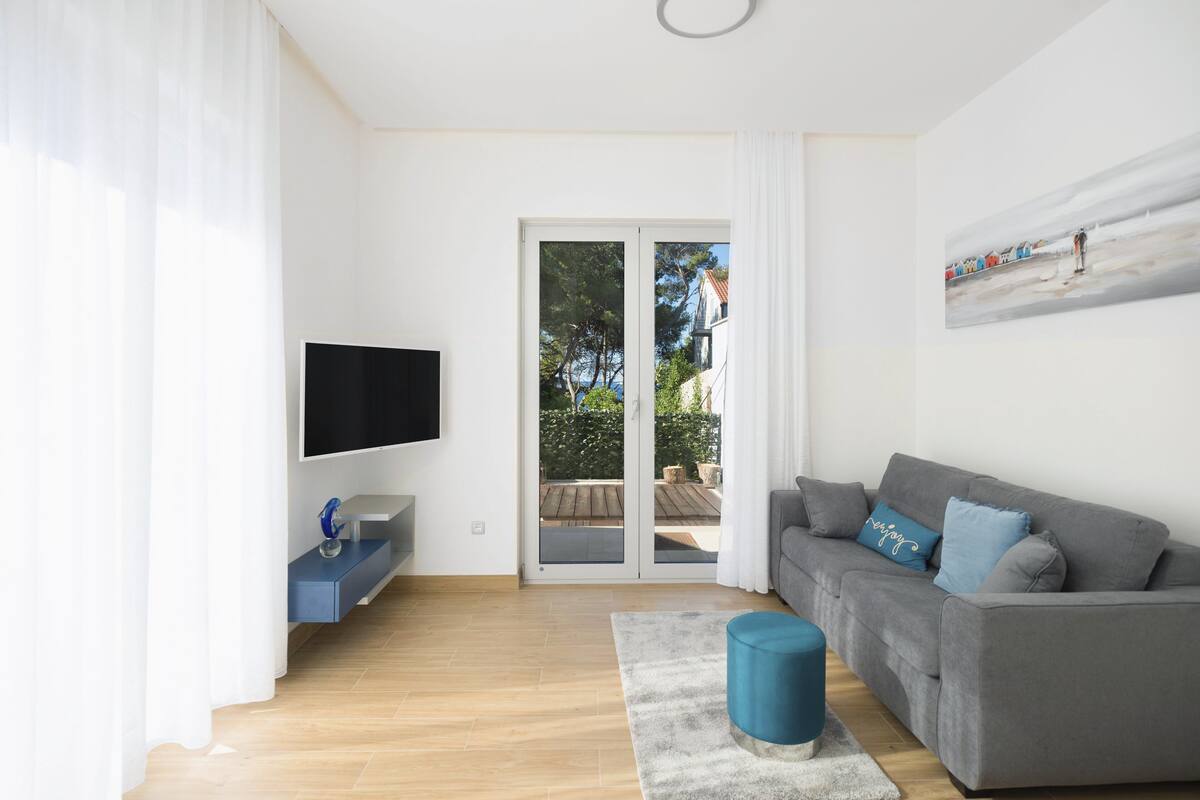 Una sala de estar moderna con un sofá gris, un televisor de pantalla plana, una mesita azul pequeña y vistas a un área exterior a través de un ventanal.