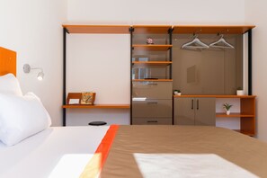 A hotel room with a bed, a wardrobe with shelves, a desk with a chair, and a small plant. - Beach Residences Caroline (Hvar)