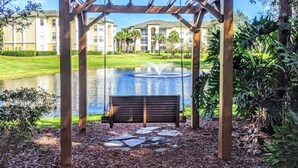 A wooden pergola with a bench in a landscaped garden area with a pond and buildings in the background. - Cosy Condo near Disney and Parks. season price. (Kissimmee)