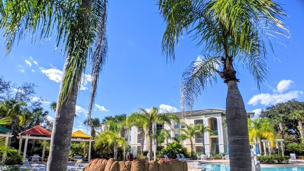 A resort pool area with palm trees, a circular water feature, and a building with a tiled roof. - Cosy Condo near Disney and Parks. season price. (Kissimmee)