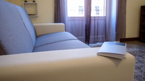 A beige sofa with a book on it, a small shelf with plants, and a window with curtains. - Housestation (Palermo)