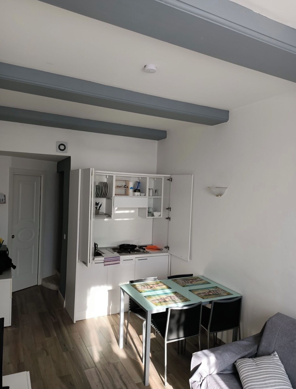 A compact kitchen with a dining area, featuring a table with chairs, a built-in cabinet with kitchenware, and a ceiling-mounted smoke detector.