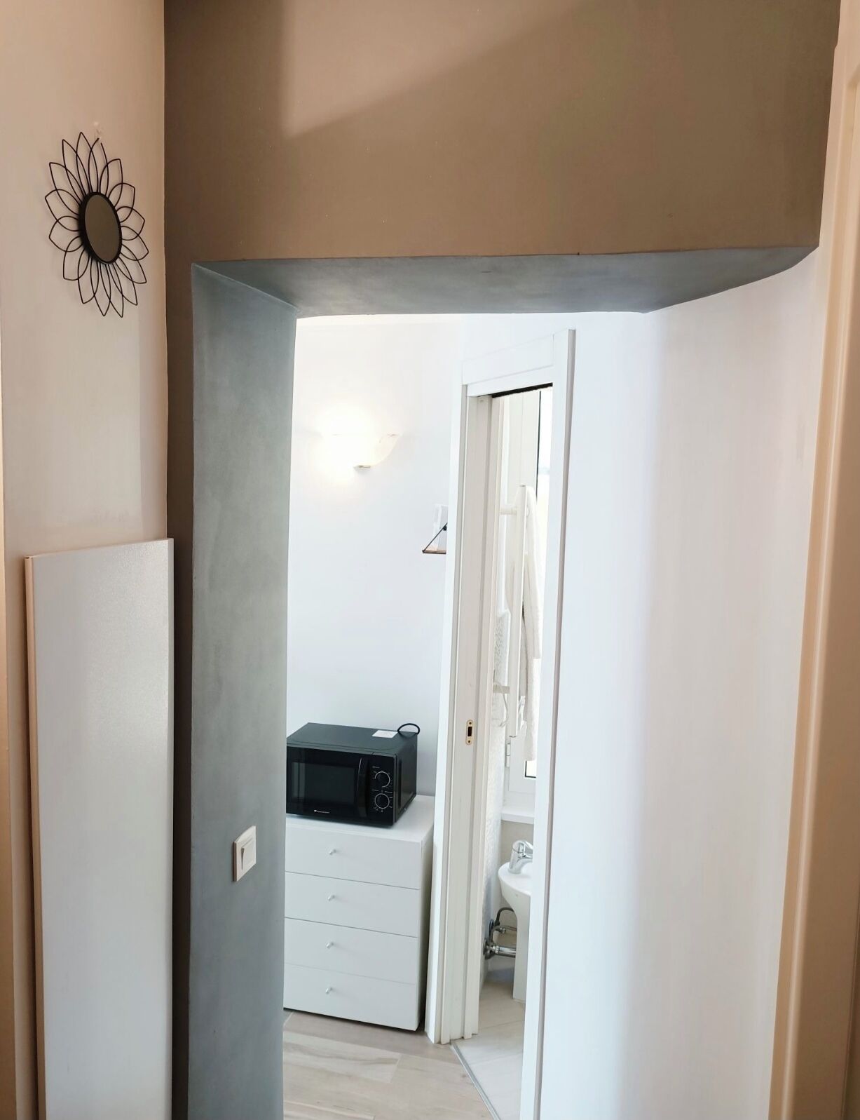 Un corridoio stretto che conduce a una stanza con un forno a microonde su un mobile bianco, una porta bianca e un punto luce fissato al muro.
