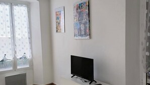 Un salon moderne avec un téléviseur à écran plat posé sur un meuble blanc, de grandes fenêtres ornées de rideaux légers et deux affiches encadrées accrochées au mur.