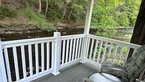 Une terrasse en bois avec une balustrade blanche donnant sur une rivière, une chaise recouverte d’un tissu à motifs floraux et une table en verre.