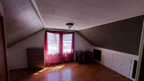 A room with a sloped ceiling, wooden flooring, a window with sheer curtains, a small cabinet, and a flat-screen TV mounted on the wall.