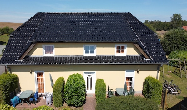 A two-story house with a dark tiled roof, a front door, and a small patio area with a table and chairs.