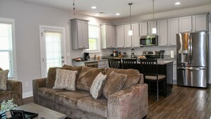 A modern kitchen with white cabinets, stainless steel appliances, and a dining area with a brown sofa and patterned cushions. - Pinehurst Golf Retreat, Pool Table, Fire Pit! (Pinehurst)