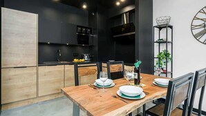 A modern kitchen with dark cabinetry, a wooden dining table set for four, and a decorative metal shelf with books and a plant. - Stylish Apartment | Parking and Close to the Old Town (Poznań)