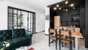 A modern living room with a dark green sofa, a dining area with a wooden table and chairs, and a kitchen with black cabinets and a hanging light fixture. - Stylish Apartment | Parking and Close to the Old Town (Poznań)