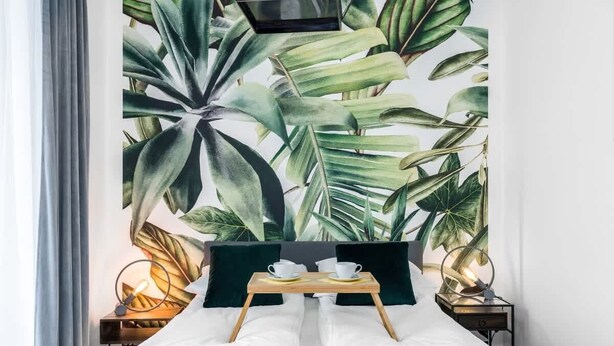A neatly made bed with white linens, a wooden bedside table with a cup and saucer, and a wall mural of tropical leaves. - Stylish Apartment | Parking and Close to the Old Town (Poznań)