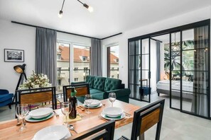 Un salón moderno con un sofá verde, mesa de comedor lista para cuatro comensales y amplias ventanas con vistas al vecindario.