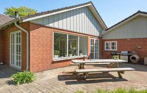 Ett hus byggt av tegel med ett stort fönster, en grill och ett träbord på baksidan av trädgården.