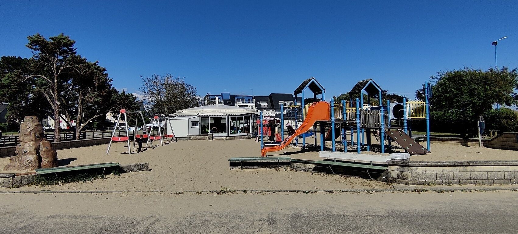 Ein Spielplatz mit Rutschen und Klettergerüsten, umgeben von Bäumen und blauem Himmel.